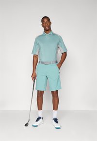 Golfový outfit pozostávajúci z svetlo tyrkysového polo trička s šedými akcentmi a zodpovedajúcich šortiek. Obsahuje šedý opasok a biele golfové topánky s námorníckymi šnúrkami.