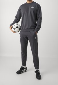 Felpa grigia e pantaloni della tuta abbinati, entrambi in un materiale morbido. La felpa presenta un logo, i pantaloni hanno tasche. Pallone da calcio nero tenuto in mano.