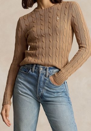 Kvinde iført en beige kabelstrikket sweater stoppet ned i lyseblå højtsiddende jeans, med den ene hånd i lommen og skulderlangt brunt hår.