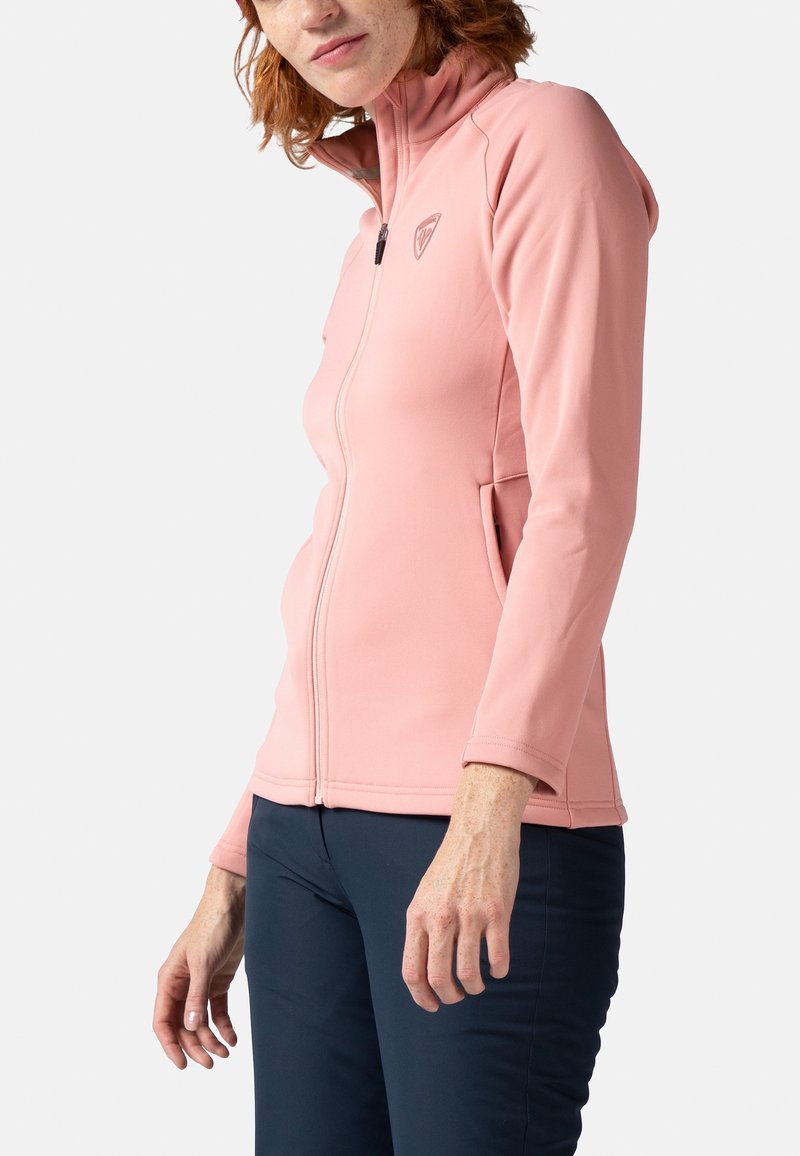Veste zippée rose clair en tissu doux et extensible, avec un col montant et deux poches latérales, associée à un pantalon bleu marine.