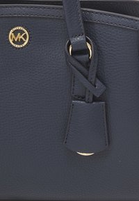 Détail d'un sac à main en cuir bleu foncé texturé avec logo Michael Kors doré et anse nouée près d'une attache anneau dorée.