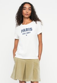 Woman with curly hair wearing a white "PARIS EST. 2007" T-shirt and beige pleated corduroy skirt, standing with hands in pockets.