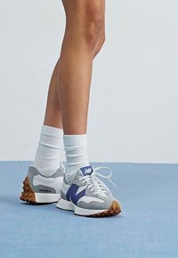 Sneaker grigie e bianche con un logo viola, dotate di una suola texturizzata e lacci, indossate con calze bianche a coste.