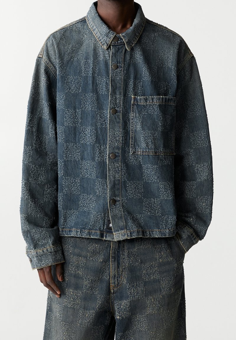 Personne portant une veste et un pantalon en denim bleu foncé avec un motif à carreaux subtil, mains dans les poches avant, visage non visible.