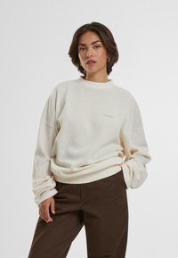 Cremefarbene Sweatshirt mit strukturiertem Strickmuster, hochgeschlossenem Kragen und gesticktem Logo, kombiniert mit dunkelbraunen Hosen.