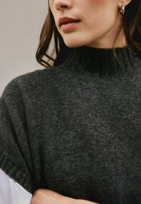 Woman wearing a dark gray knit turtleneck sweater with loose brown hair and a small hoop earring visible on her left ear.