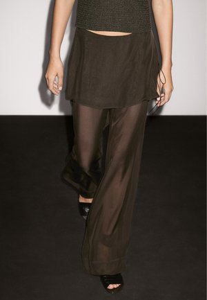 Un mannequin défile dans une tenue olive semi-transparente composée d'un haut court et de pantalons superposés, fluides, associés à des talons ouverts noirs sur un fond sombre.