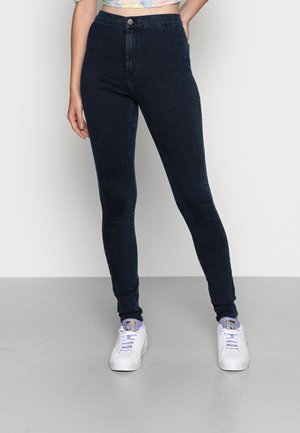 Person iført højtaljede, mørkeblå skinny jeans og hvide sneakers med lyse lilla snørebånd, stående mod en ensfarvet baggrund.