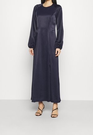 Kobieta ubrana w granatową satynową suknię maxi z długim rękawem i okrągłym dekoltem oraz czarne sandały na paski, stojąca na tle jednolitego jasnego tła.