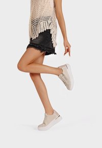 Sneakers in pelle beige con punta arrotondata e suola bianca, caratterizzati da dettagli dorati e una finitura dei lacci testurizzata. Indossati con una gonna di jeans nera.