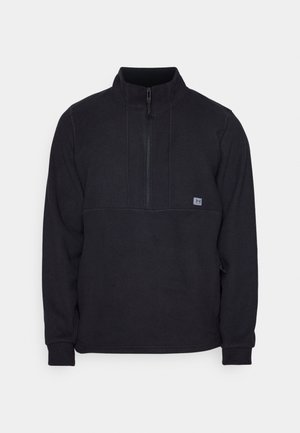 Črn flis pulover s pol-zadrgo in detajlom šiva na prsih, ki vključuje majhen logotip na levi strani. Mehka tekstura, dolgi rokavi.