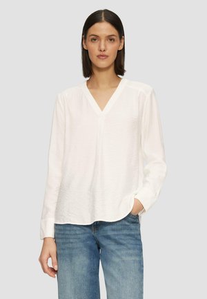 Blouse blanche à manches longues avec un décolleté en V, coupe décontractée et tissu texturé. Une paire de jeans bleus complète l'ensemble.