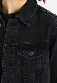 Black denim jacket featuring a pointed collar, two chest pockets with decorative stitching, and silver button accents. Textured fabric.