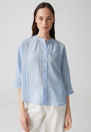 Hellblaue Bluse mit Rundhalsausschnitt, dreiviertel Ärmeln und vorderem Knopfverschluss. Hergestellt aus einem transparenten, leichten Stoff.