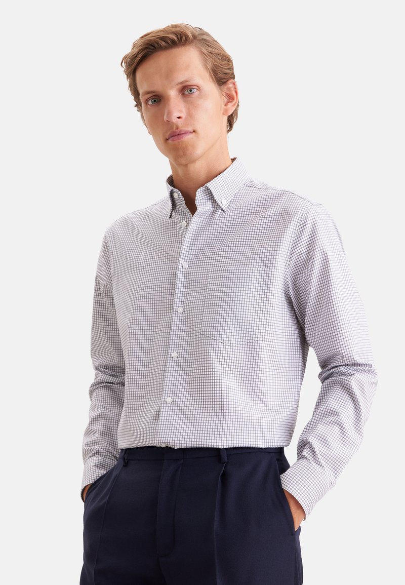 Jeune homme portant une chemise blanche à petits carreaux et un pantalon bleu foncé, debout les mains dans les poches.