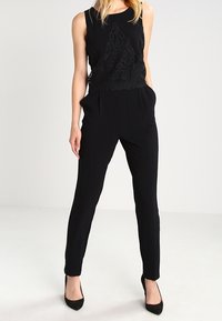 Mono negro con un diseño sin mangas y detalles de encaje en el bodice. Las piernas estrechas y los bolsillos mejoran la funcionalidad.