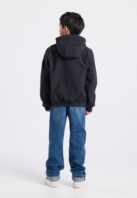 Mustapaitainen huppari, jossa on sileä pinta, yhdistettynä sinisiin farkkuihin. Leikkaus on rento, näkyvillä saumoilla ja slight tapered -lahkeilla.