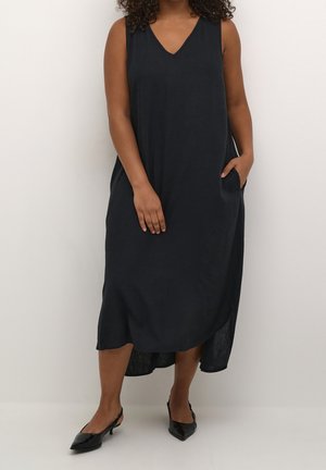 Femme portant une robe midi noire sans manches avec des poches et des escarpins slingback noirs à bout pointu, debout devant un fond uni.