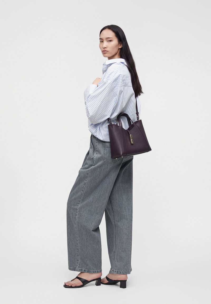 Femme aux longs cheveux foncés portant une chemise ample rayée, un jean large gris, des sandales noires à talons, portant un sac à main violet foncé sur l'épaule.