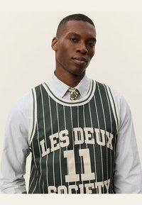 Grøn stribet basketballtrøje med "LES DEUX 11 SOCIETY" trykt i hvidt. Båret over en lyseblå stribet skjorte og mønstrede slips.