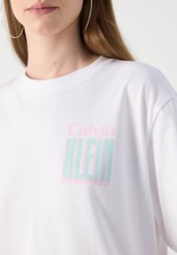 T-shirt en coton blanc avec le logo "Calvin KLEIN" en rose pastel et vert menthe, avec un col rond et une coupe décontractée.