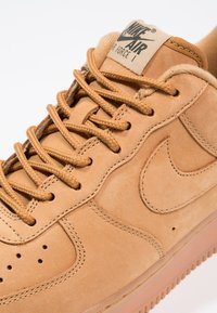 Tan leather Nike Air Force 1 kenkä, jossa on pyöreät beige nauhat, rei'itetty varvas, ommeltu yksityiskohtia ja kumipohja.