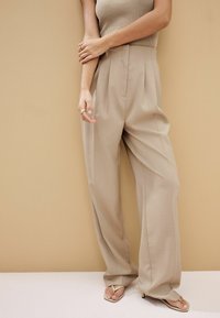 Pantalones beige con plisados, fabricados en una tela suave, que presentan una cintura alta y piernas anchas y fluidas, combinados con sandalias minimalistas de punta abierta.