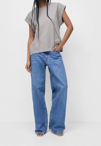 Grå bomulds t-shirt med rund hals og korte ærmer, parret med lyseblå, vide jeans. Modellen har hånden i lommen og står foran en neutral baggrund.