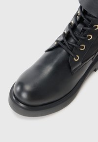 Black leather ankle boot with a rounded toe, gold eyelets, and black laces. Features a textured finish and a sturdy black rubber sole.