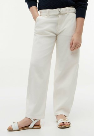 Persona che indossa pantaloni bianchi a vita alta e gamba larga con cintura coordinata e sandali bianchi open-toe con dettagli dorati, in piedi su sfondo chiaro.