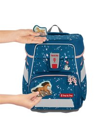 Zaino blu con motivo floreale, mano che attacca una toppa a forma di cavallo. Presenta accenti rosa, strisce riflettenti e una parte inferiore grigia.