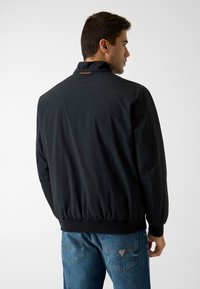 Veste bomber noire avec col montant, poignets et ourlet côtelés, et finition textile lisse. Présente un détail de logo discret au dos.