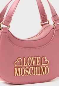 Rožainā Love Moschino soma ar zeltainu logotipu, ar rāvējslēdzēju aizdari un dubultām rokturiem ar zeltainiem gredzeniem, attēlota tuvplānā.