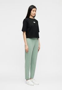 Γυναίκα που στέκεται, φοράει μαύρο κοντό μπλουζάκι, ανοιχτοπράσινο φαρδύ παντελόνι και λευκά αθλητικά παπούτσια, σε απλό φωτεινό φόντο.