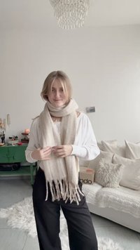 Jasnobeżowy szal z frędzlami na krawędziach, wykonany z miękkiego materiału, luźno układający się wokół szyi, noszony na białej koszuli z długim rękawem.