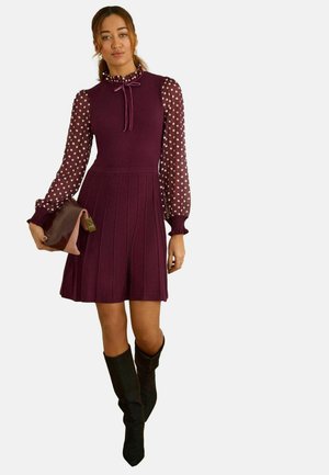 Lila ruha testhezálló felsőrésszel, fodros gallérral és pöttyös, átlátszó ujjakkal. Barna kézitáskával és fekete, térdig érő csizmával kombinálva.