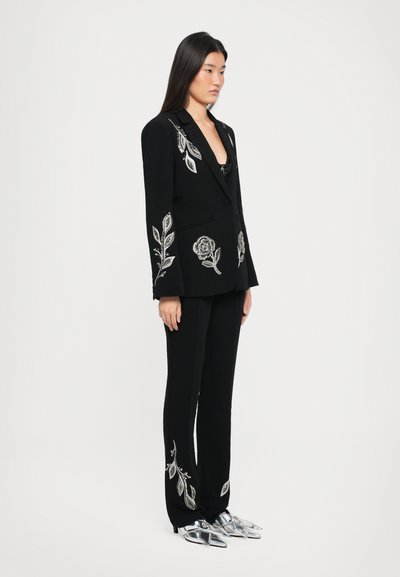 Black tailored suit with silver floral embroidery on the blazer and pants. The model wears metallic shoes. Simple background with a neutral tone.