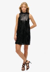 Vestido negro sin mangas adornado con lentejuelas, con un escote alto y una silueta acampanada. Combinado con tacones negros de tiras.