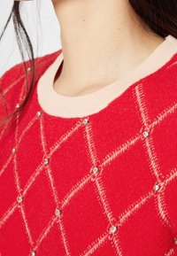Acercamiento a un suéter rojo con un escote beige claro, que presenta un patrón de diamantes adornado con pequeñas cuentas de cristal.
