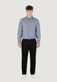 Camicia azzurra a maniche lunghe con una texture sottile, abbinata a pantaloni neri e sneakers bianche. La camicia ha un colletto classico e una fila di bottoni.
