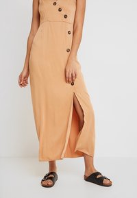 Robe sans manches couleur beige avec des boutons en diagonale et une fente latérale, portée avec des sandales noires à boucle sur un fond gris clair.