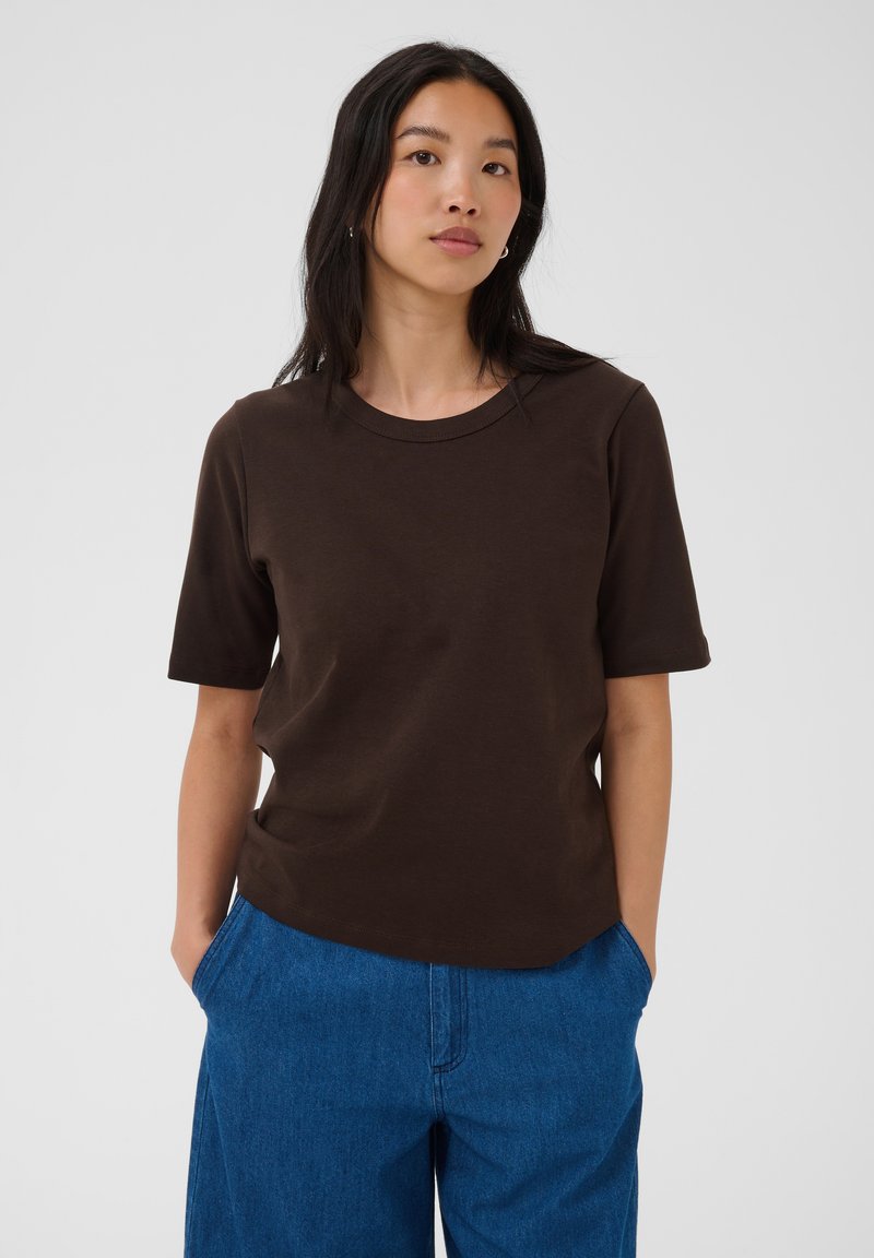 Jeune femme aux longs cheveux foncés portant une chemise marron à manches courtes et un jean bleu, debout les mains dans les poches sur un fond uni.