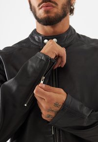 Veste en cuir noir avec un col haut et des boutons-pression en argent. Dotée de poignets zippés et d'une texture lisse. Des mains tiennent la fermeture éclair.