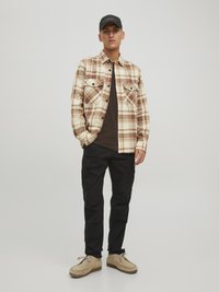Kariertes Flanellhemd in Beige und Braun, schwarze Cargo-Hosen und beige Schuhe mit Schnürsenkeln. Hut mit Text auf der Vorderseite.