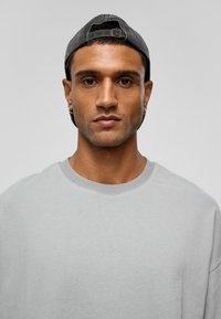 Lichtgrijze sweatshirt met een brede ronde hals, gedragen door een persoon met een zwarte baseballpet. Glad textuur en een ontspannen fit.
