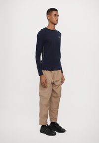 Jūrinės tamsiai mėlynos spalvos ilgomis rankovėmis marškiniai su auksiniais siūlų akcentais, derinami su šviesiai rudos spalvos krovinių kelnėmis. Juodi sportbačiai baigia aprangą.