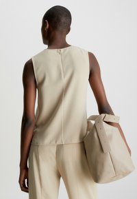 Beige ärmlös topp med dragkedja i ryggen, kombinerad med en matchande beige väska med bred rem och subtil textur.