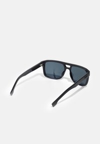 Occhiali da sole neri con forma rettangolare, lenti grigio scuro, montatura in plastica lucida e aste sottili con un branding discreto.