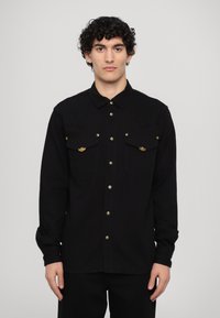 Camisa negra de botones con dos bolsillos de parche, herrajes en tono dorado, mangas largas y cuello clásico. Fabricada con un tejido texturizado.