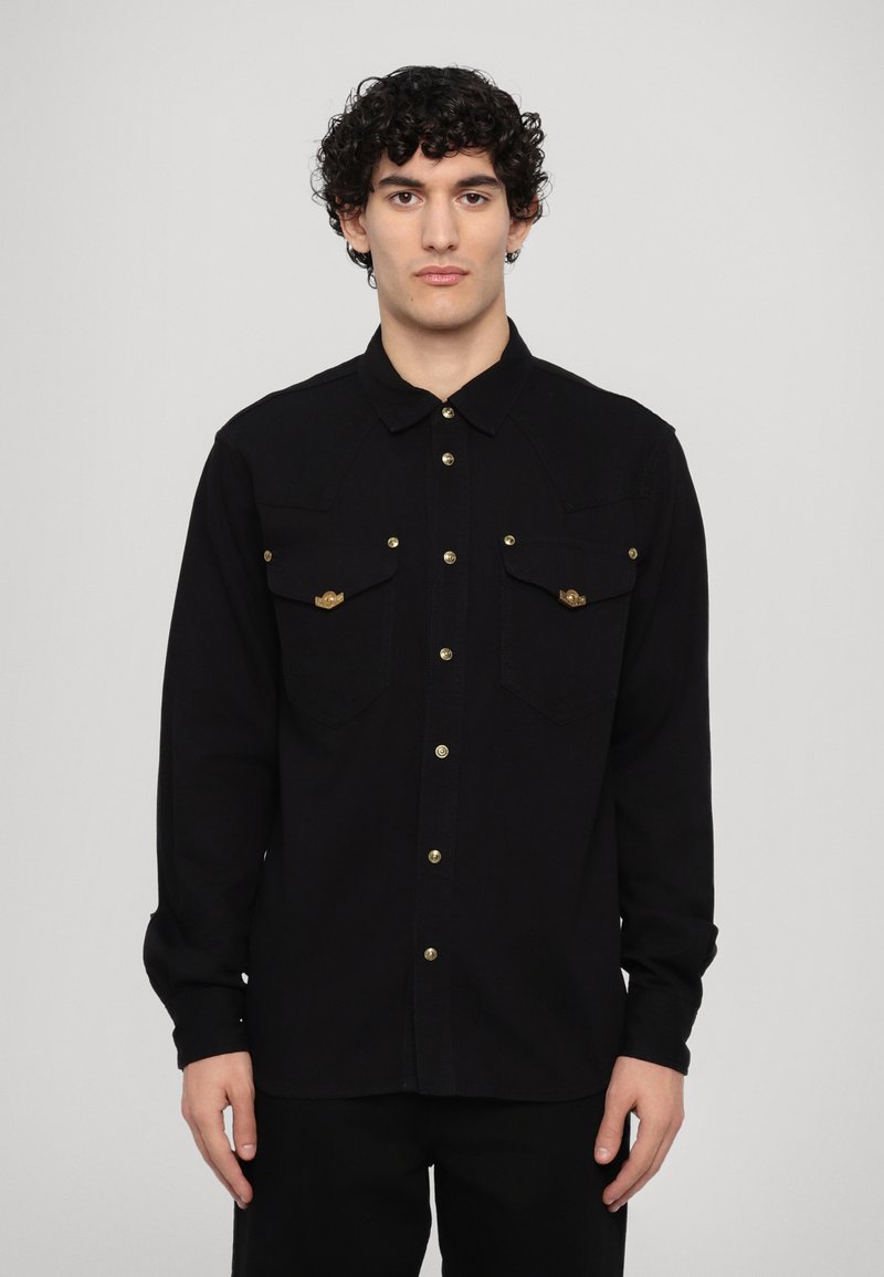 Camisa negra de botones con dos bolsillos de parche, herrajes en tono dorado, mangas largas y cuello clásico. Fabricada con un tejido texturizado.